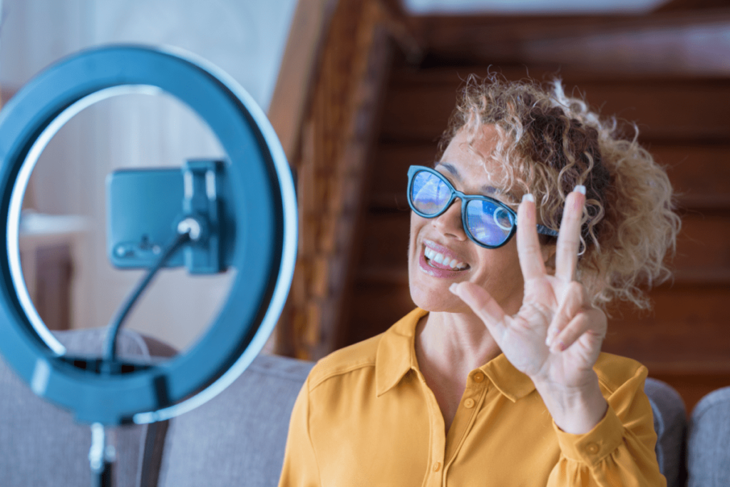 A young lady with the pace sign sitting in front of a cellphone and an attached ring light.