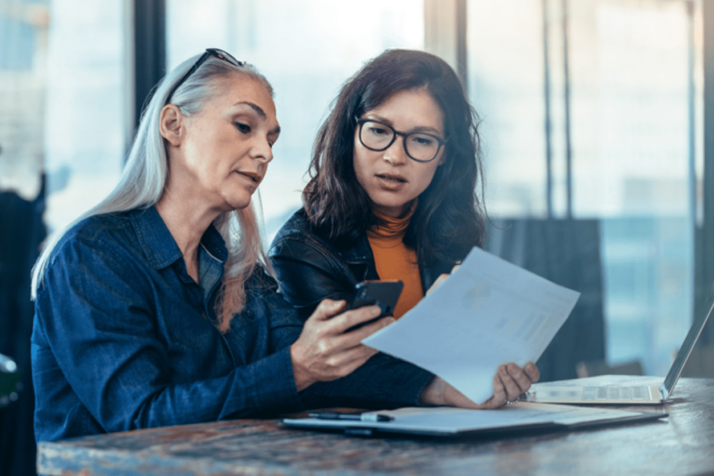 Two female social media managers comparing page performance data on a cell phone and printed analytics report